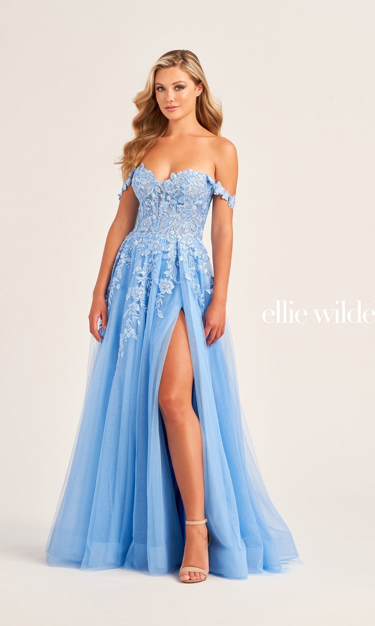 A woman poses elegantly in the Ellie Wilde Off-Shoulder Long Prom Dress EW35101, featuring a light blue hue, thigh-high slit, and lace detailing against a plain background.